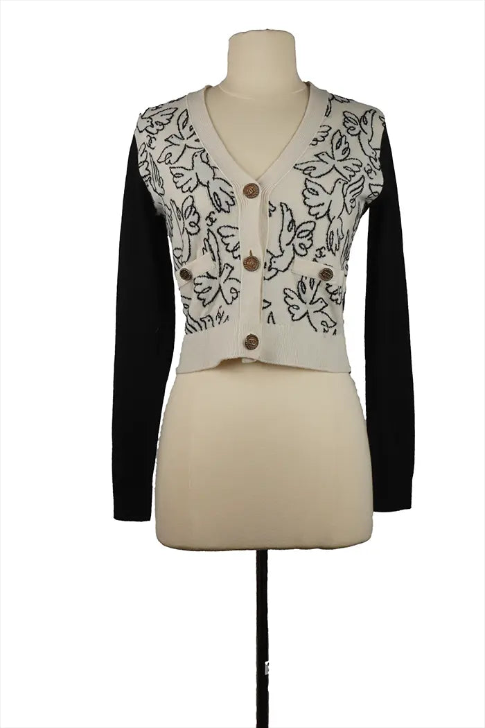 Cárdigan Chanel Croisière 2022 en maniquí, parte delantera de cachemira blanca adornada con palomas negras, cuello de pico, botón CC dorado, mangas negras.